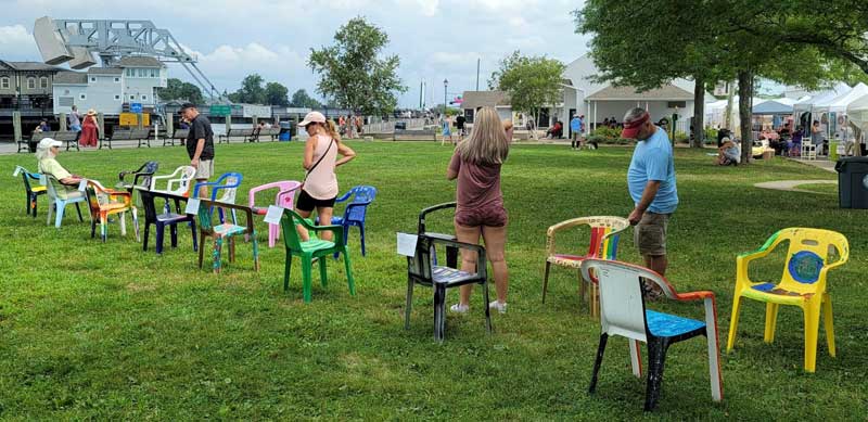 A photograph of the Conversation Chairs installation in River Park during the 2021 Mystic Outdoor Art Festival. 16 chairs face each other in two long rows as visitors read their descriptions. 